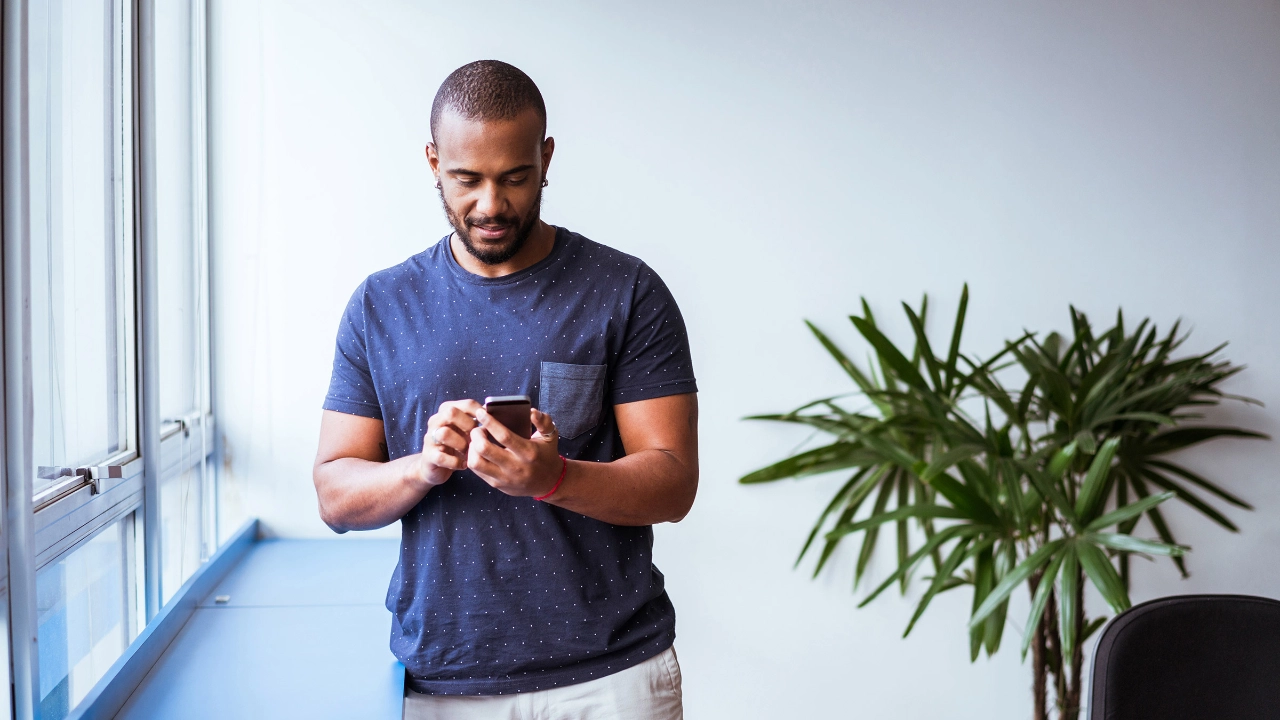 Ein dunkelhaariger man steht vor einem Fesnster und tippt auf seinem Smartphone.
