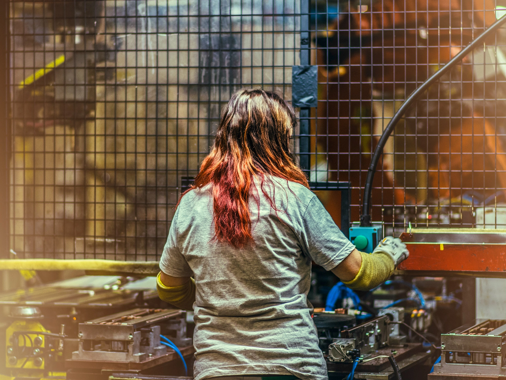 Die Rückansicht einer Frau mit langen bunten Haaren, die an einer technischen Anlage arbeitet und dabei Arbeitskleidung und Schutzhandschuhe trägt.